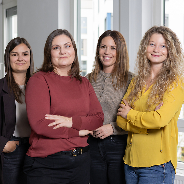 Vier junge Frauen, nebeneinander stehend vor Fenster, lächeln in die Kamera