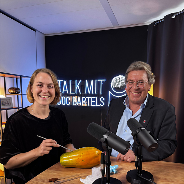 Dr. Alicia Baier und Dr. Andreas Bartels bei "Talk mit Doc Bartels" Junge Frau links mit Skalpell und älterer Mann rechts, sitzend am Studiotisch, darauf eine Papaya