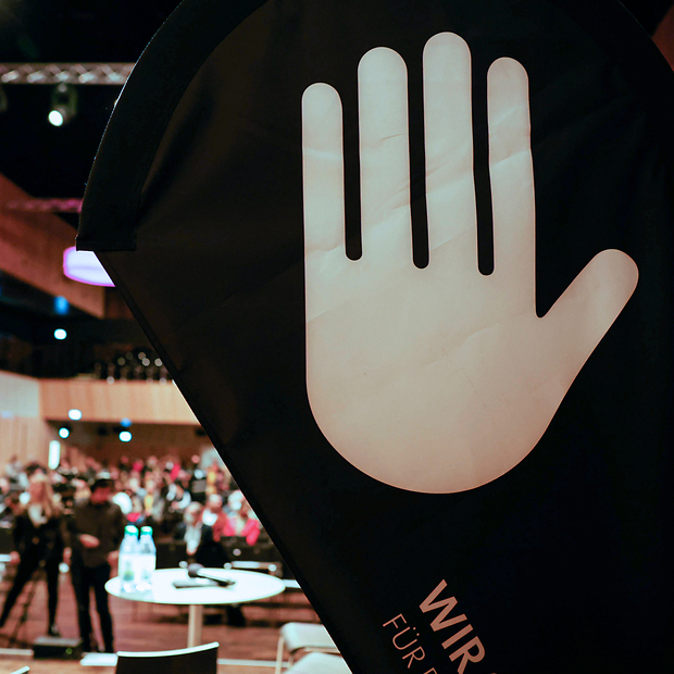 WIR SEHEN SCHWARZ Grafik von erhobener Hand auf Rollup, im Hintergrund Halle innen mit Menschen