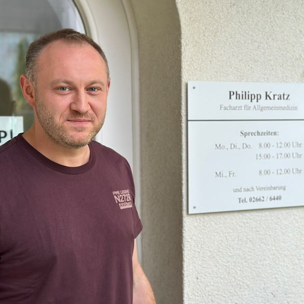 Mann mittleren Alters, lichtes Haar, im T-Shirt vor seiner Praxis, rechts daneben das Praxisschild