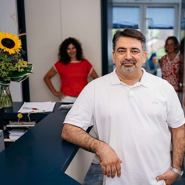 Alireza Nasserabadi, kurze Haare und Bart, weißes Poloshirt, lehnt am Empfangstresen der Praxis