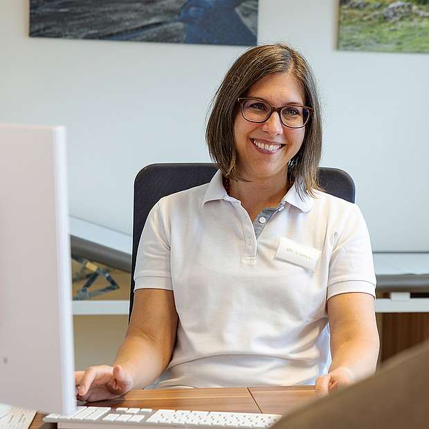 Frau mittleren Alters, Bob-Frisur, Brille, Poloshirt, lächelnd, sitzt am Schreibtisch in Praxis