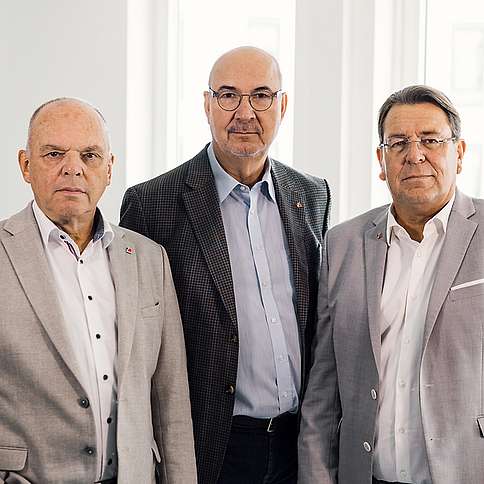 Vorstand der KV RLP 3 ältere Herren in Anzug, nebeneinander stehend, blicken ernst in die Kamera.
