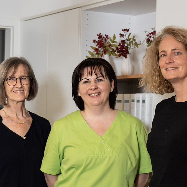 3 Frauen mittleren Alters, alle mit mittellangen Haaren, in legerer und Praxiskleidung, lächelnd