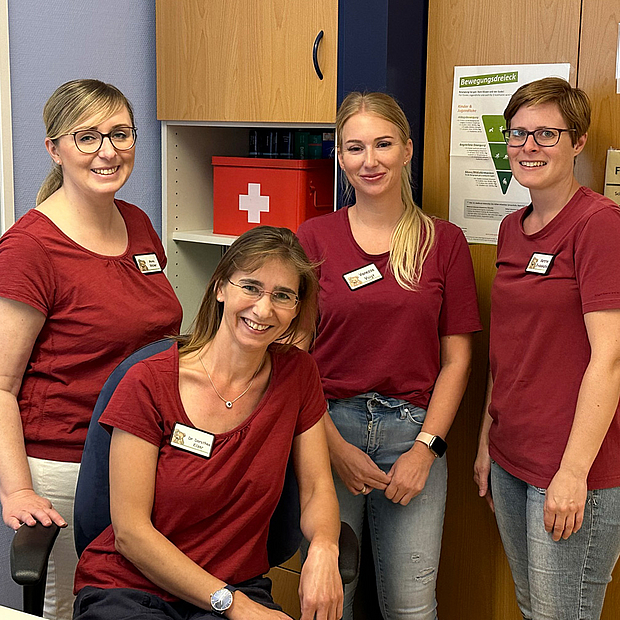 4 Frauen jungen und mittleren Alters in Teamkleidung, lächelnd, sitzen und stehen vor Schrankwand