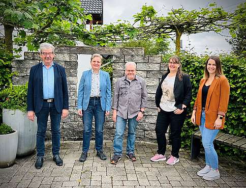 3 Frauen und Männer mittleren und höheren Alters stehen vor einer Mauer dahinter Bäume und ein Haus