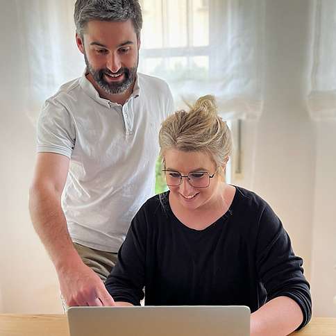 Frau und Mann mittleren Alters, sie mit Brille und Dutt sitzend am Laptop, er stehend. Sie lächeln.